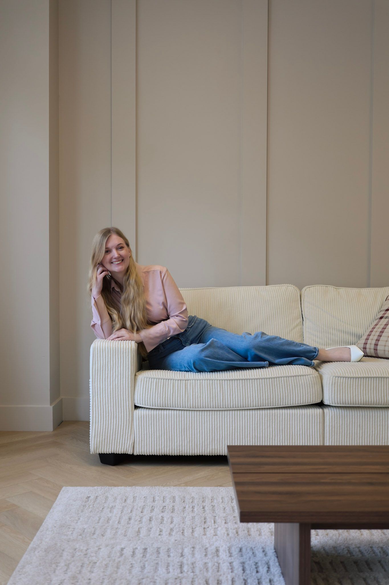 Smiling blonde woman relaxing on a cream corduroy sofa, talking on phone, wearing pink blazer and jeans in a modern living room.