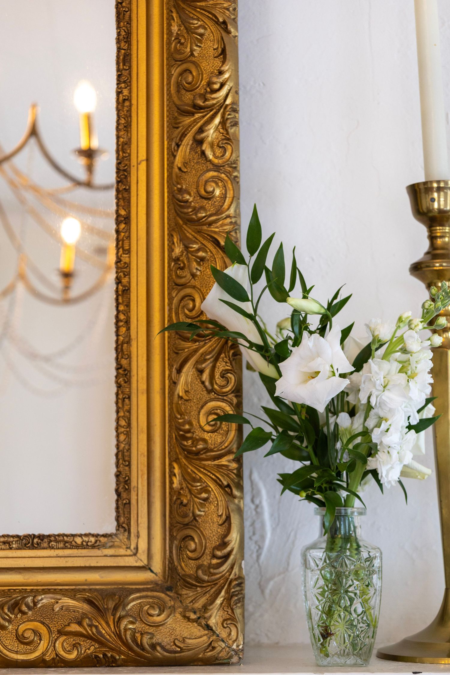 Ornate gold mirror frame beside white flowers in glass vase with brass candlestick on white mantel.