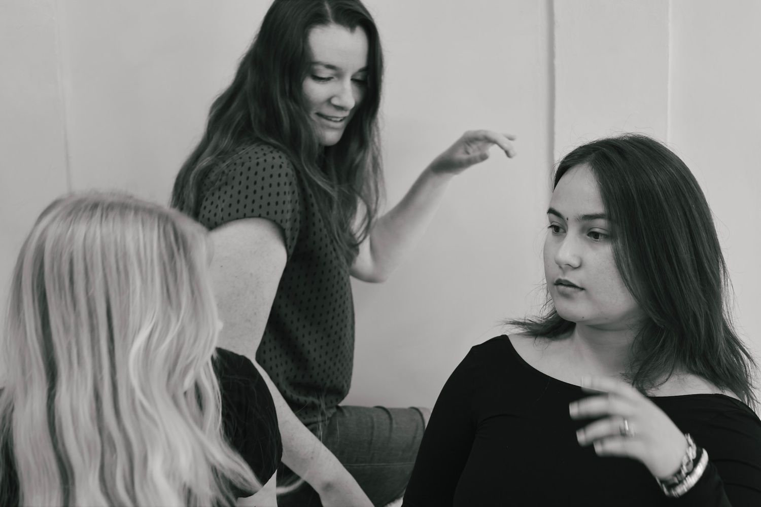 Three women conversing in black and white photo, one standing and gesturing while two others listen attentively.