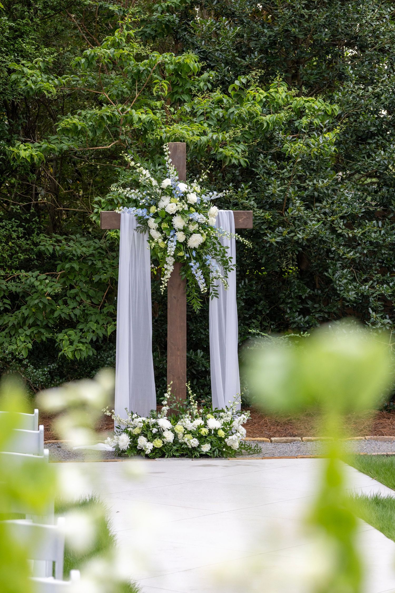 Outdoor wedding ceremony cross decorated with white floral arrangements and draped fabric, set against lush green trees.