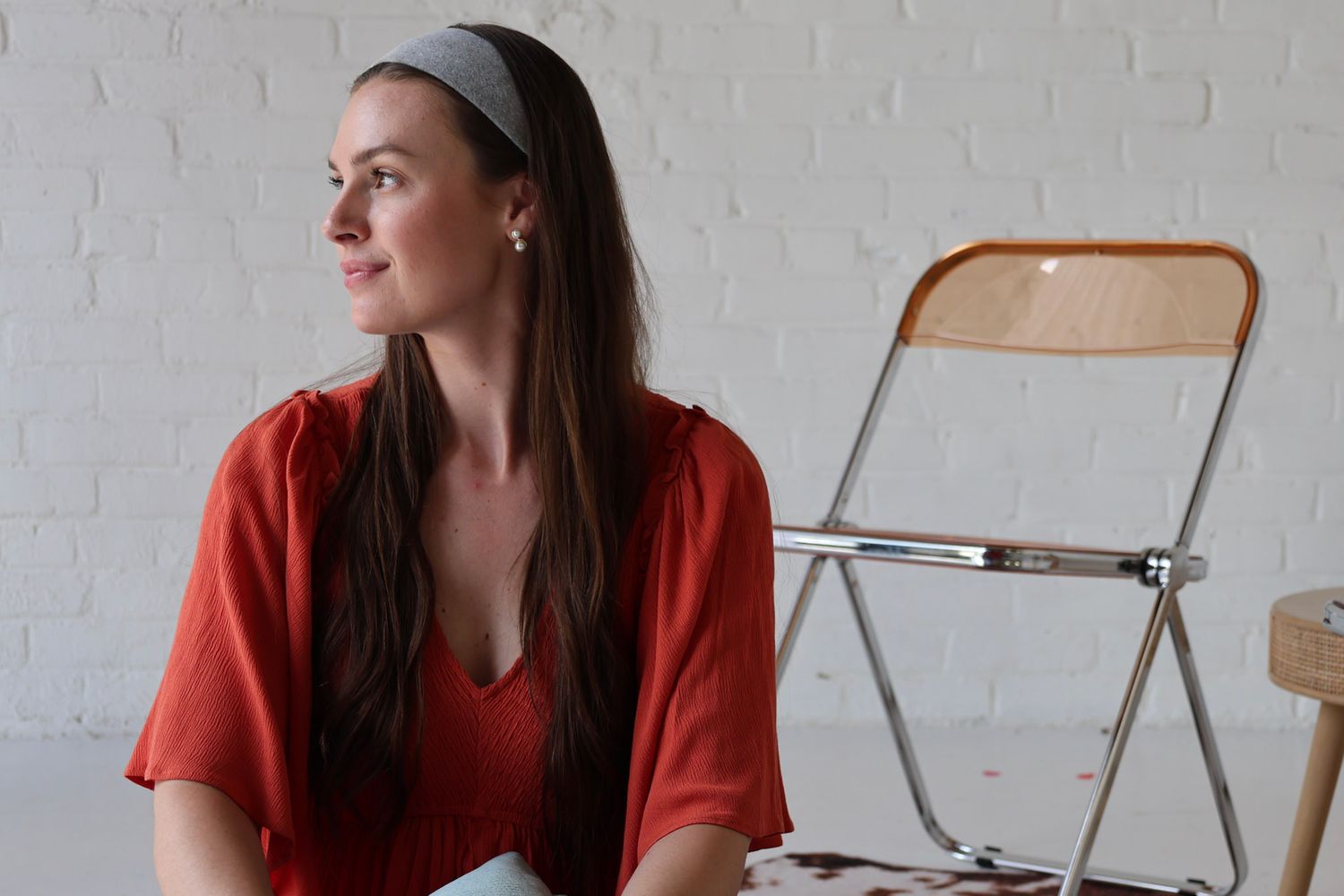 Woman in red top with gray headband sits thoughtfully beside a folding chair against a white brick wall.