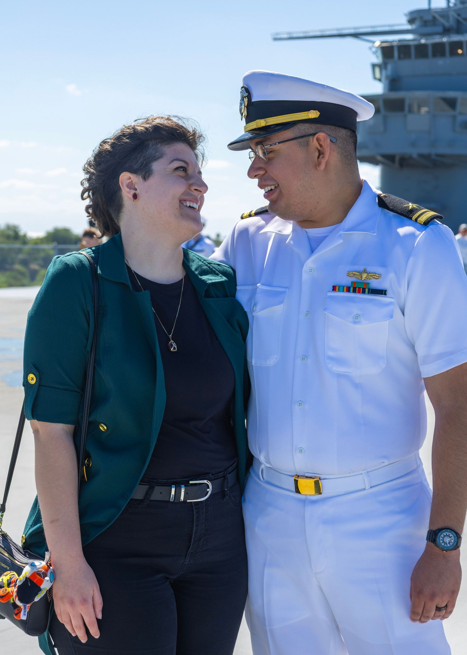 Navy officer in white uniform smiling with a woman in green jacket at a military base.