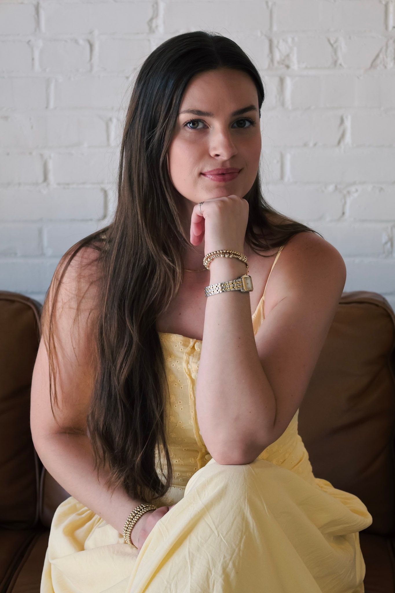 Young woman with long dark hair in yellow sundress sits on brown leather sofa, resting chin on hand, gold jewelry.