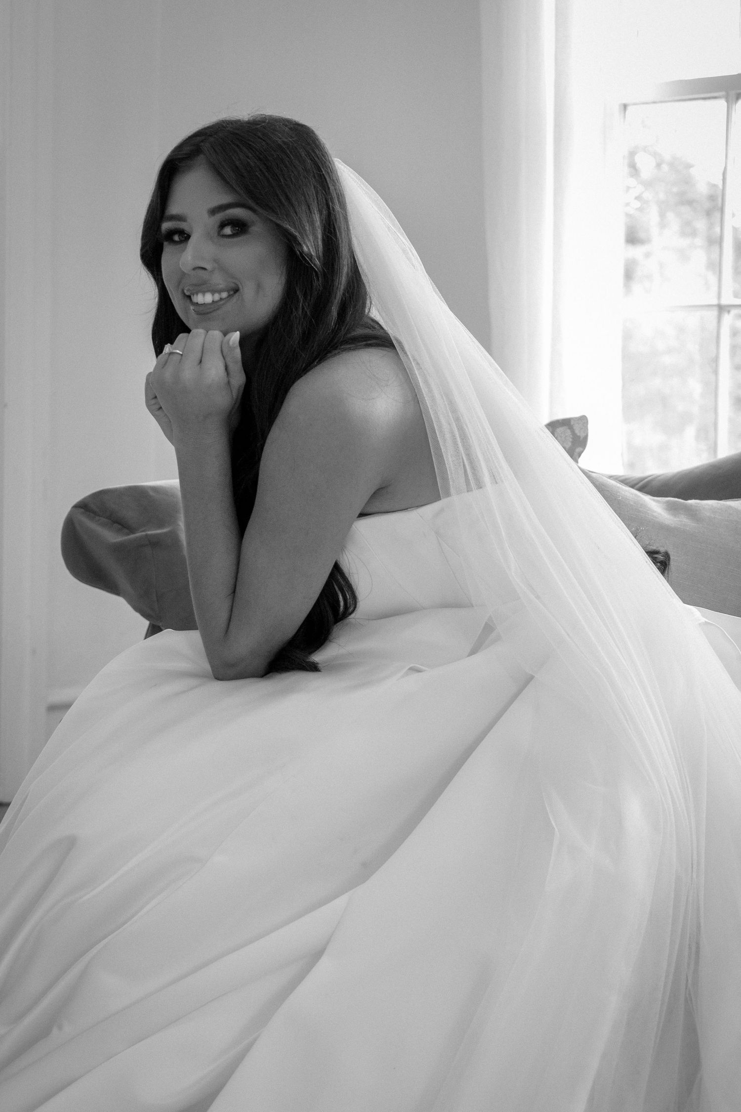 Black and white photo of a smiling bride in a strapless gown and veil, posing elegantly indoors.
