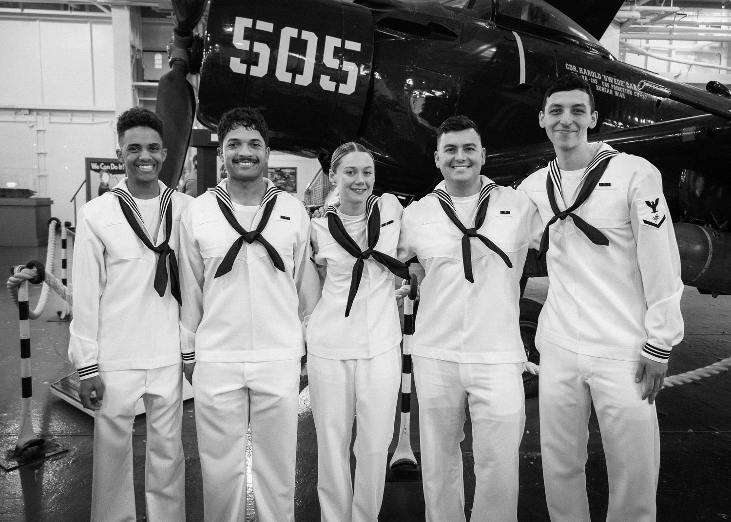 Five Navy sailors in white uniforms smile together in front of a military aircraft marked 505 in a hangar.