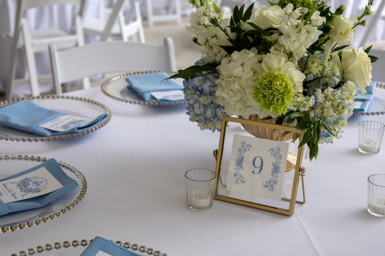 Elegant wedding reception table with blue hydrangea floral centerpiece, gold table number 9, and blue napkins on white linens.
