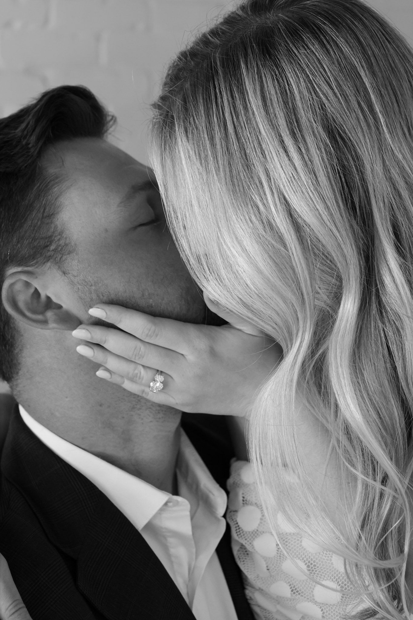 Black and white photo of a couple sharing a romantic kiss, woman displaying a diamond engagement ring on his cheek.