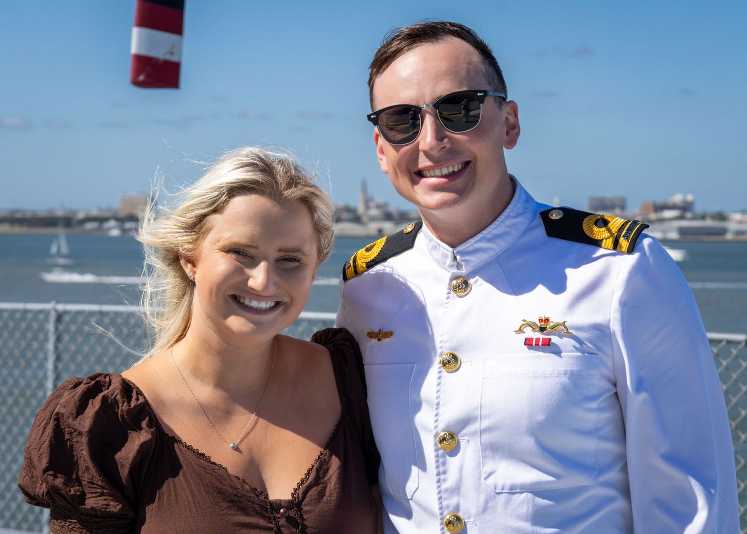 A smiling couple poses outdoors, man in white naval uniform with sunglasses, woman in brown dress, blue sky background.