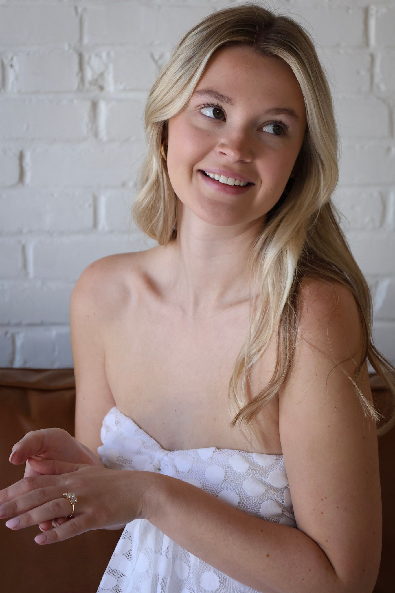 Smiling blonde bride in a white polka dot strapless dress, posing indoors against a white brick wall.