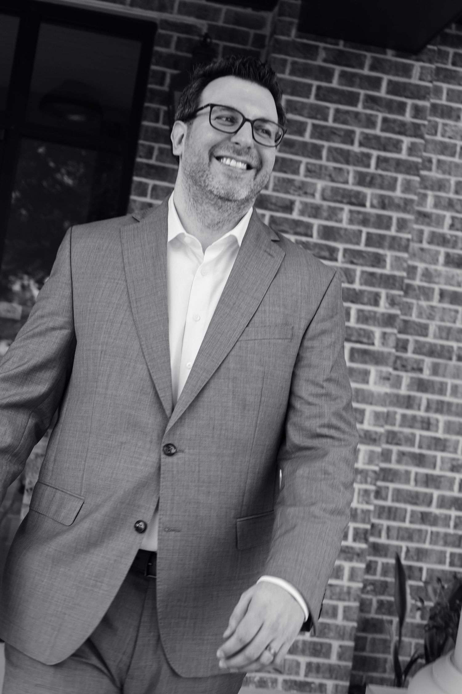 A smiling man in a gray suit and glasses stands confidently in front of a brick wall in a black and white photo.