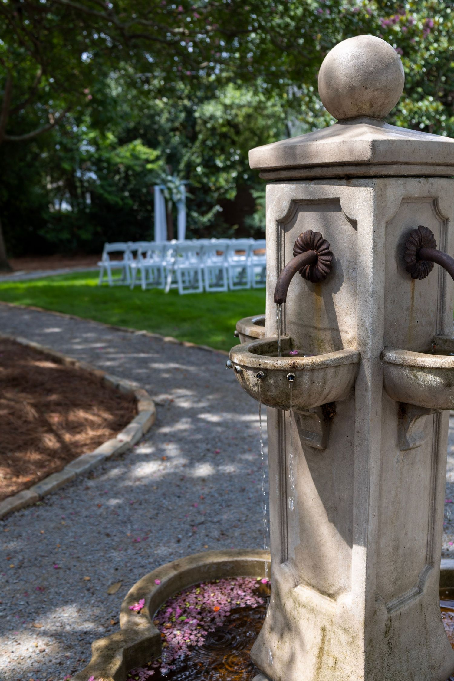 Stone garden fountain with ornate spigots in foreground, rows of white chairs set up for outdoor wedding ceremony in background.