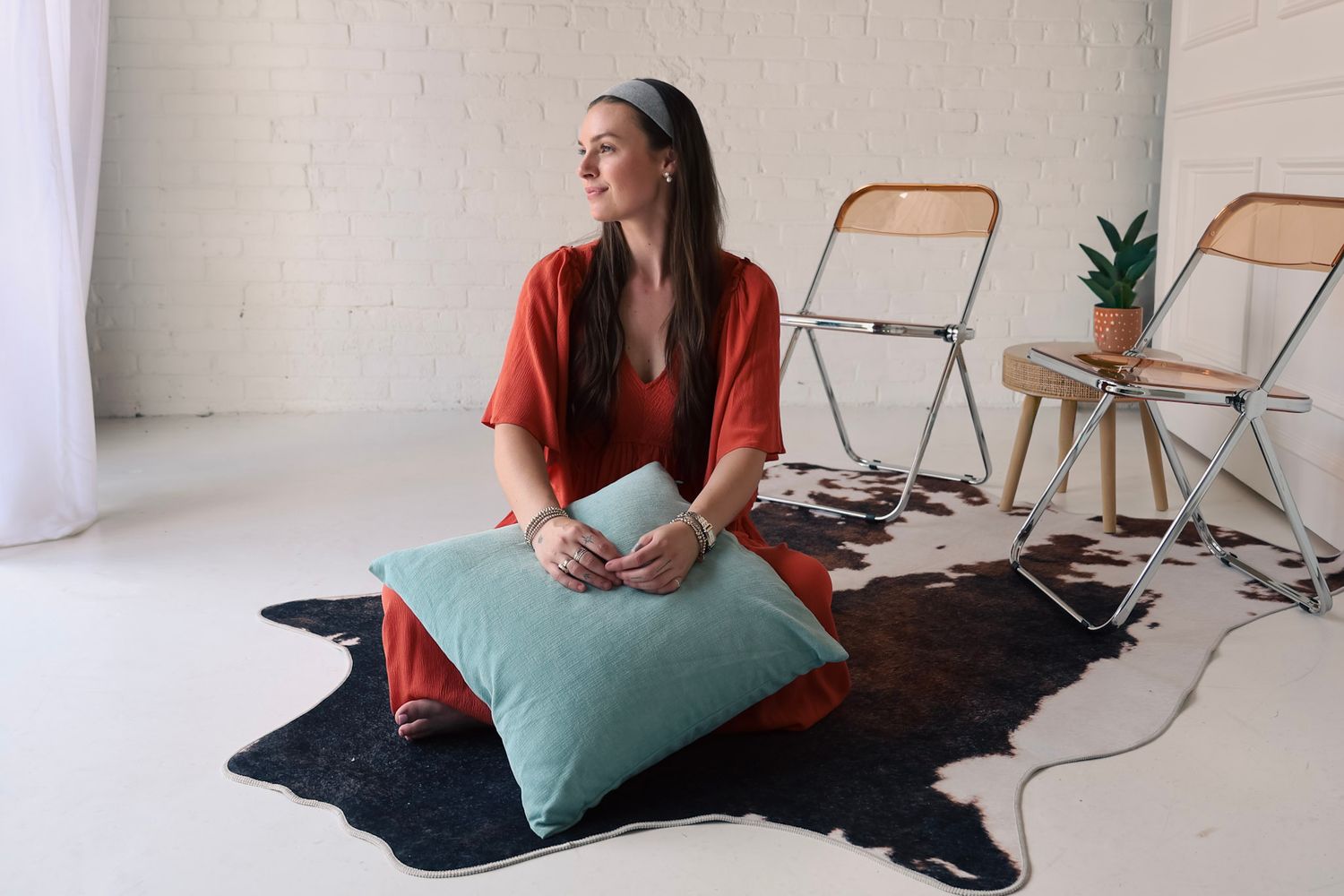 Woman in red outfit sitting on cowhide rug with teal pillow in bright white studio space.