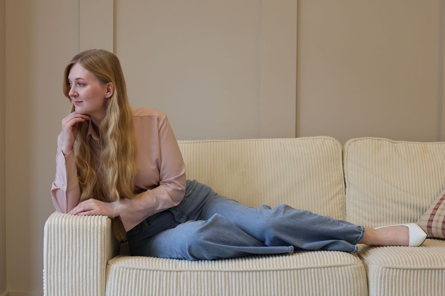 Young woman in blue jeans and pink blouse reclining thoughtfully on a cream sofa in a minimalist room.