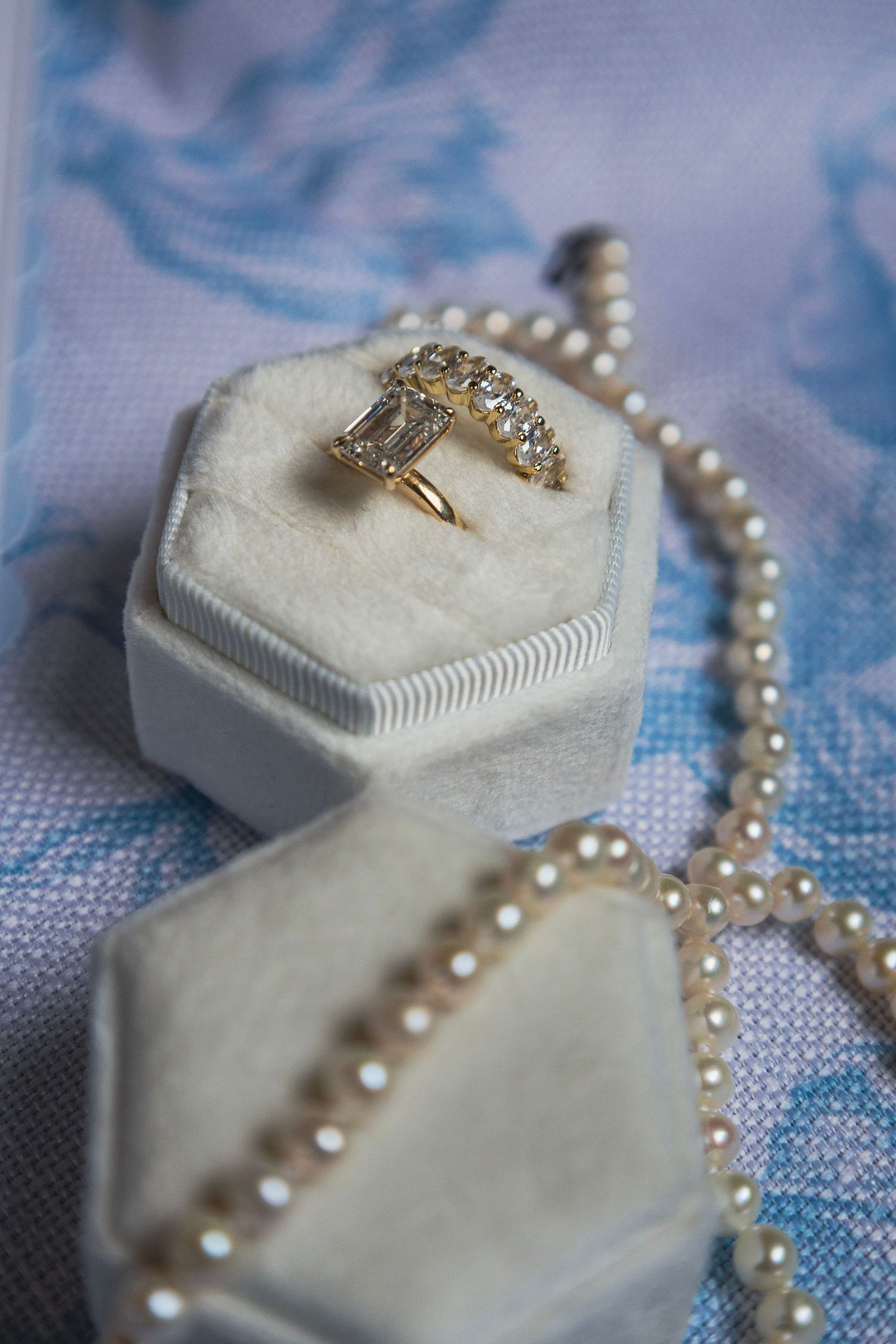 Gold diamond ring in white velvet hexagon box with pearl necklace on blue floral fabric background.