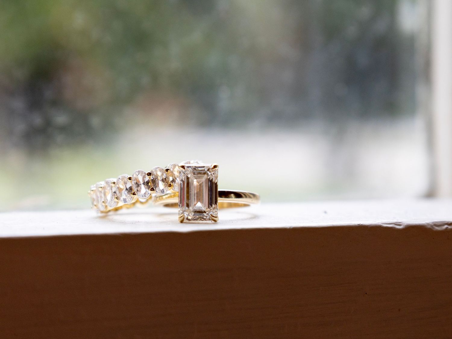 Gold engagement ring with emerald-cut morganite stone and diamond band displayed on a windowsill.