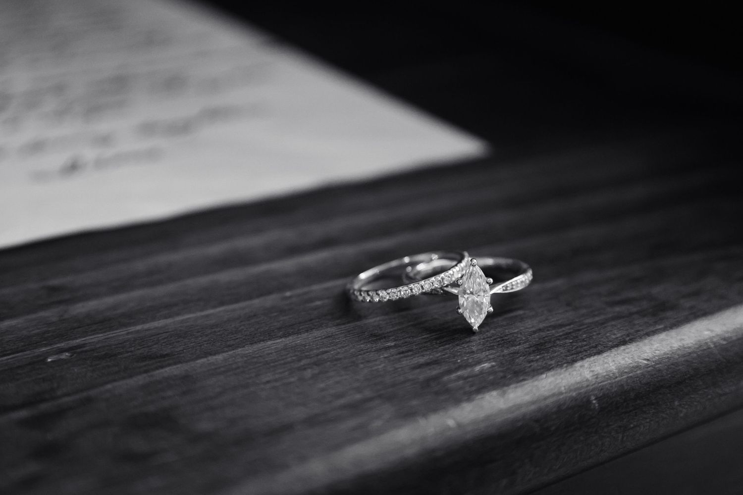 Black and white photo of a diamond marquise engagement ring on a wooden surface with a handwritten love letter in the background.
