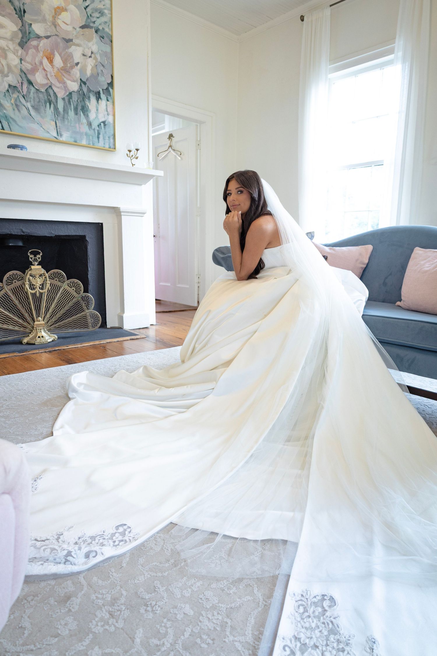 Bride in a stunning white ball gown with long veil sits elegantly in a bright, classic living room with fireplace.