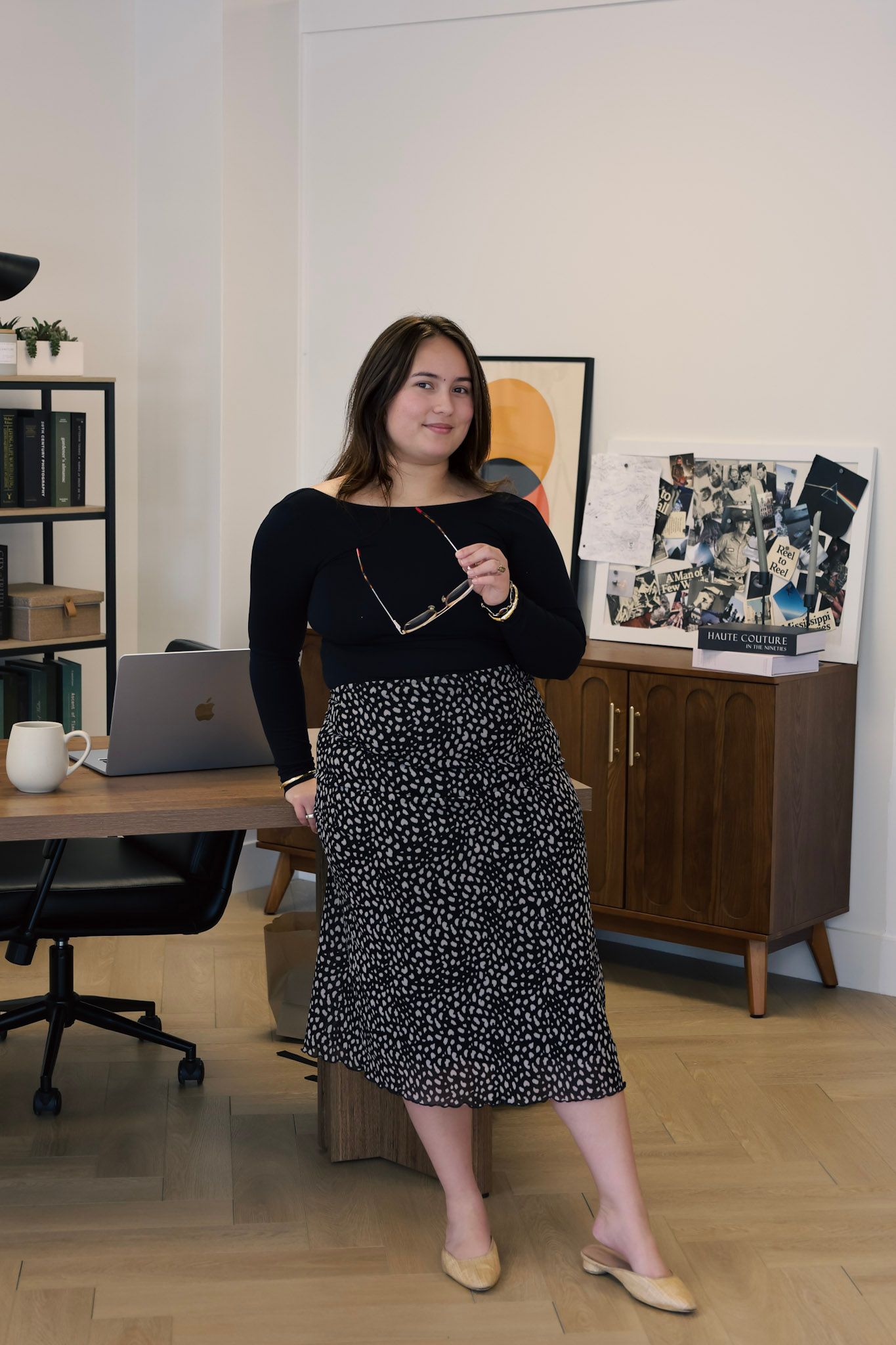 Woman in black top and floral midi skirt holding glasses, standing in a modern office space with wooden furniture.