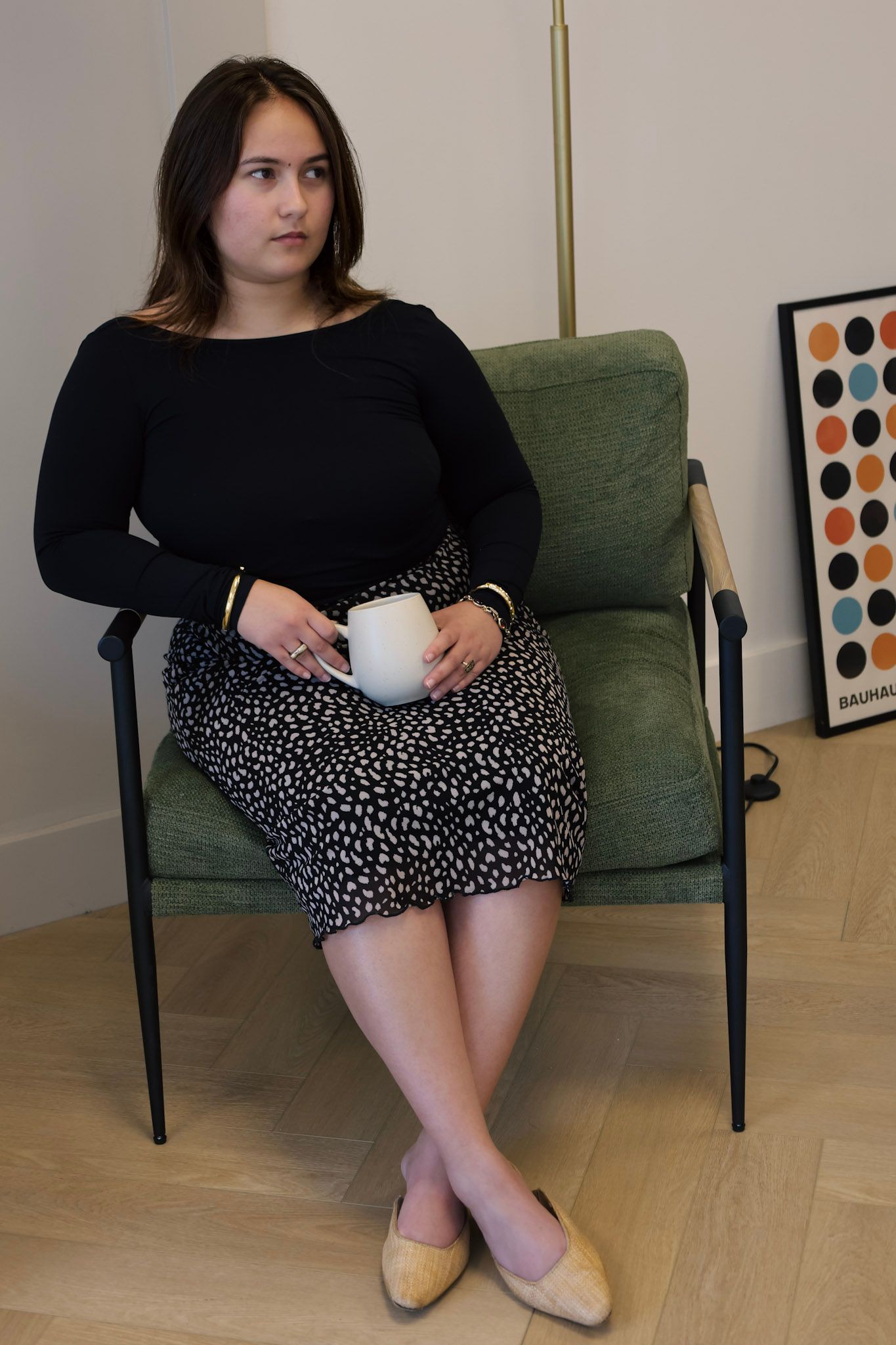 Woman in black top and spotted skirt sits in green chair holding white mug, beside colorful wall art.