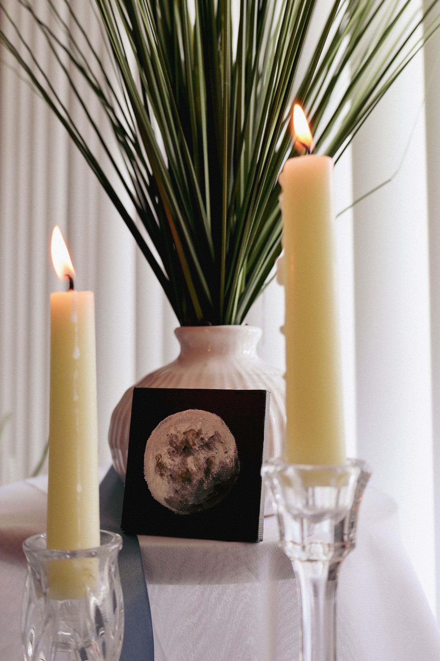 Two lit ivory taper candles in crystal holders beside a vase with palm leaves and moon art print.