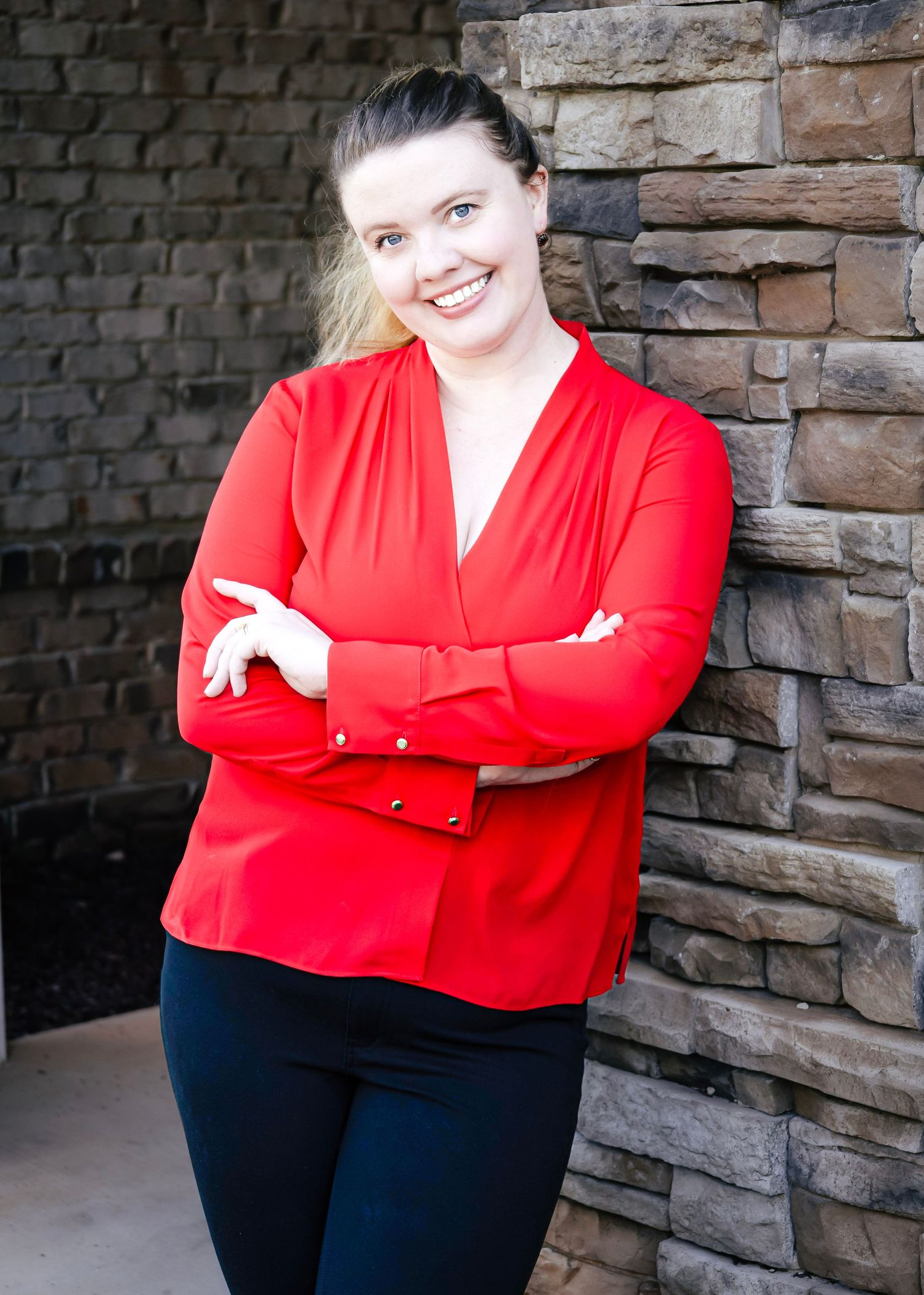A smiling woman in a red blouse with arms crossed, leaning against a stone wall in a professional headshot.