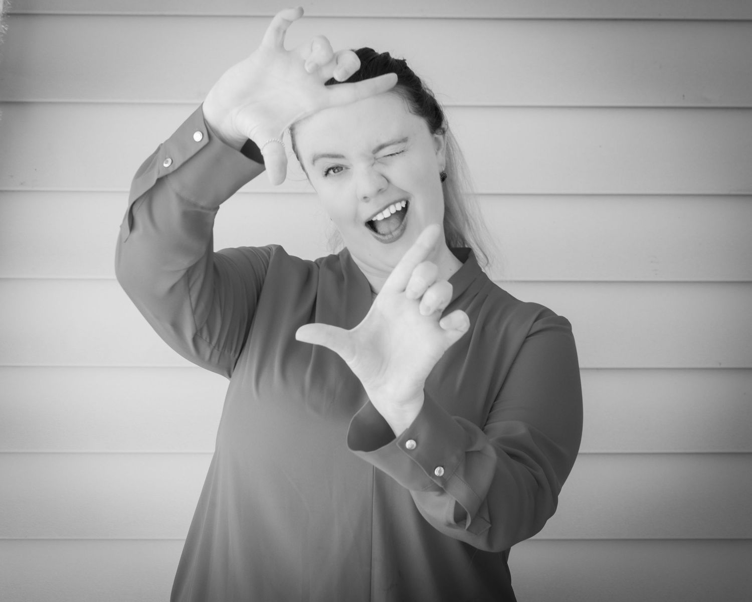 Black and white photo of a young woman making a finger frame gesture, smiling playfully at the camera.