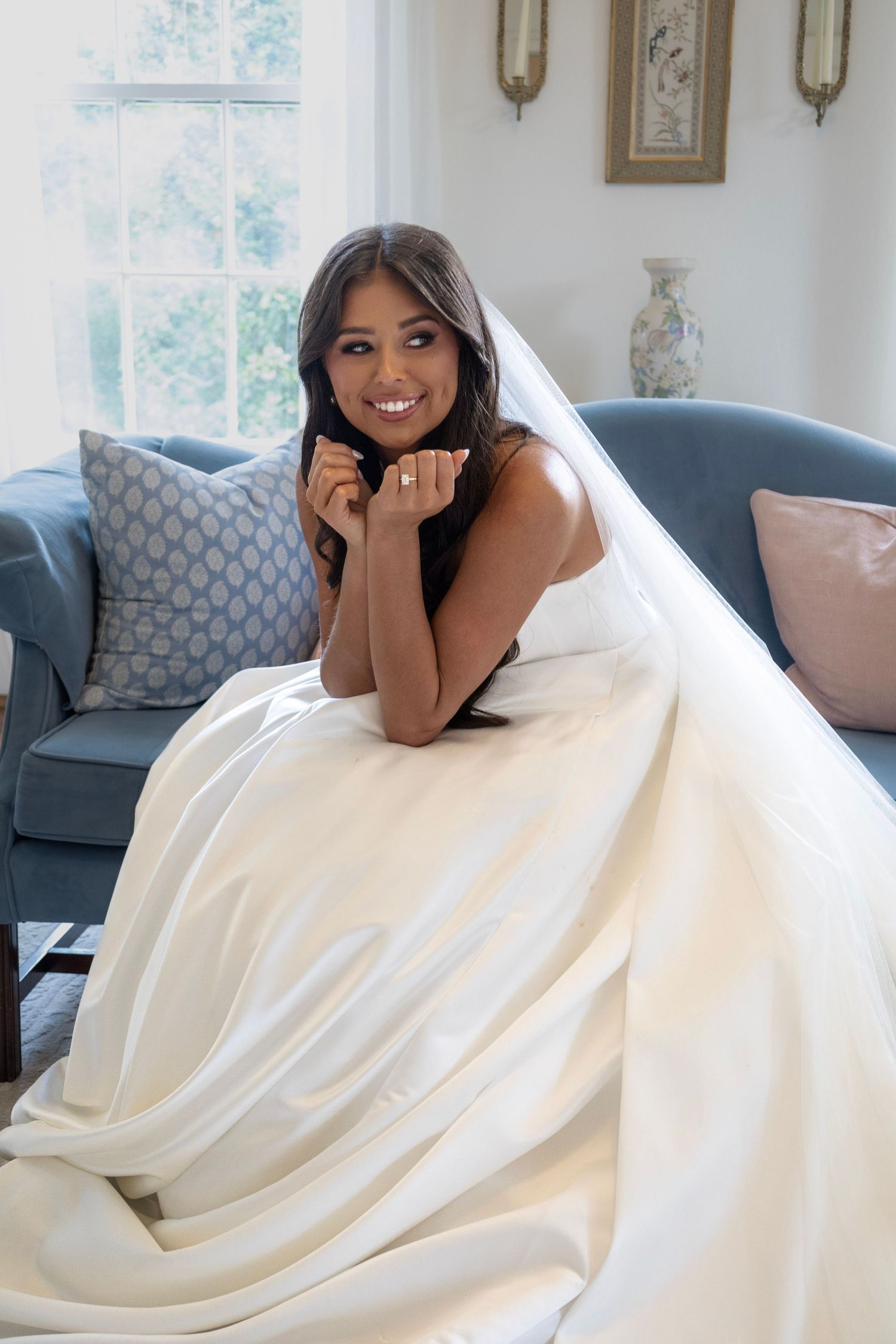 Smiling bride in white wedding gown and veil sits on blue sofa, resting chin on hands near window.