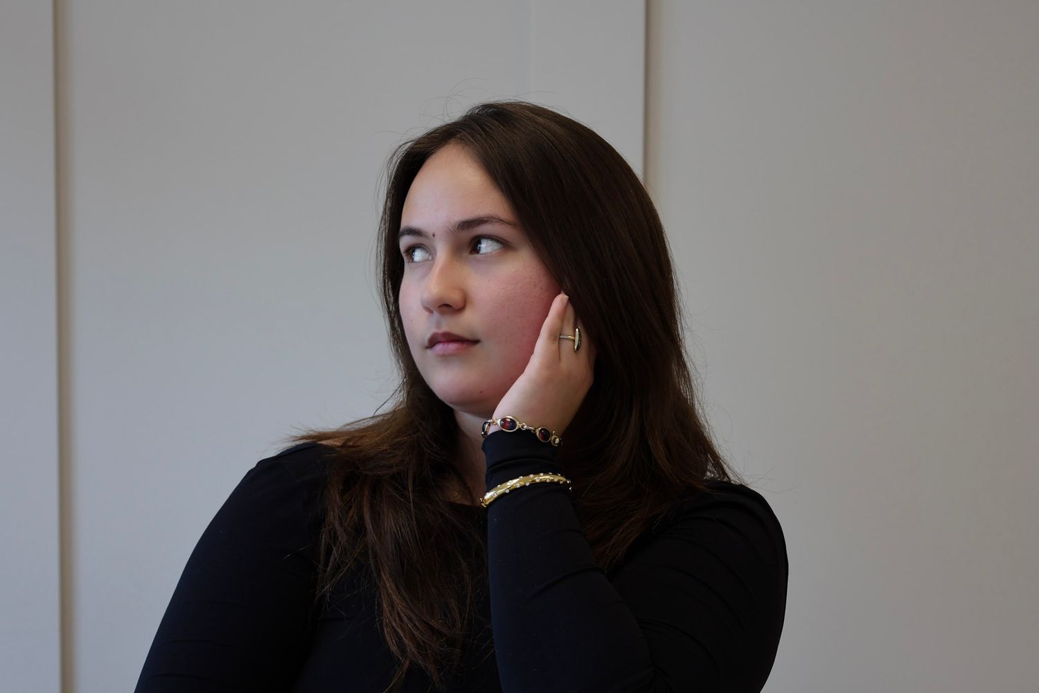 Young woman with long brown hair in black turtleneck, resting hand on chin, looking thoughtfully to the side.