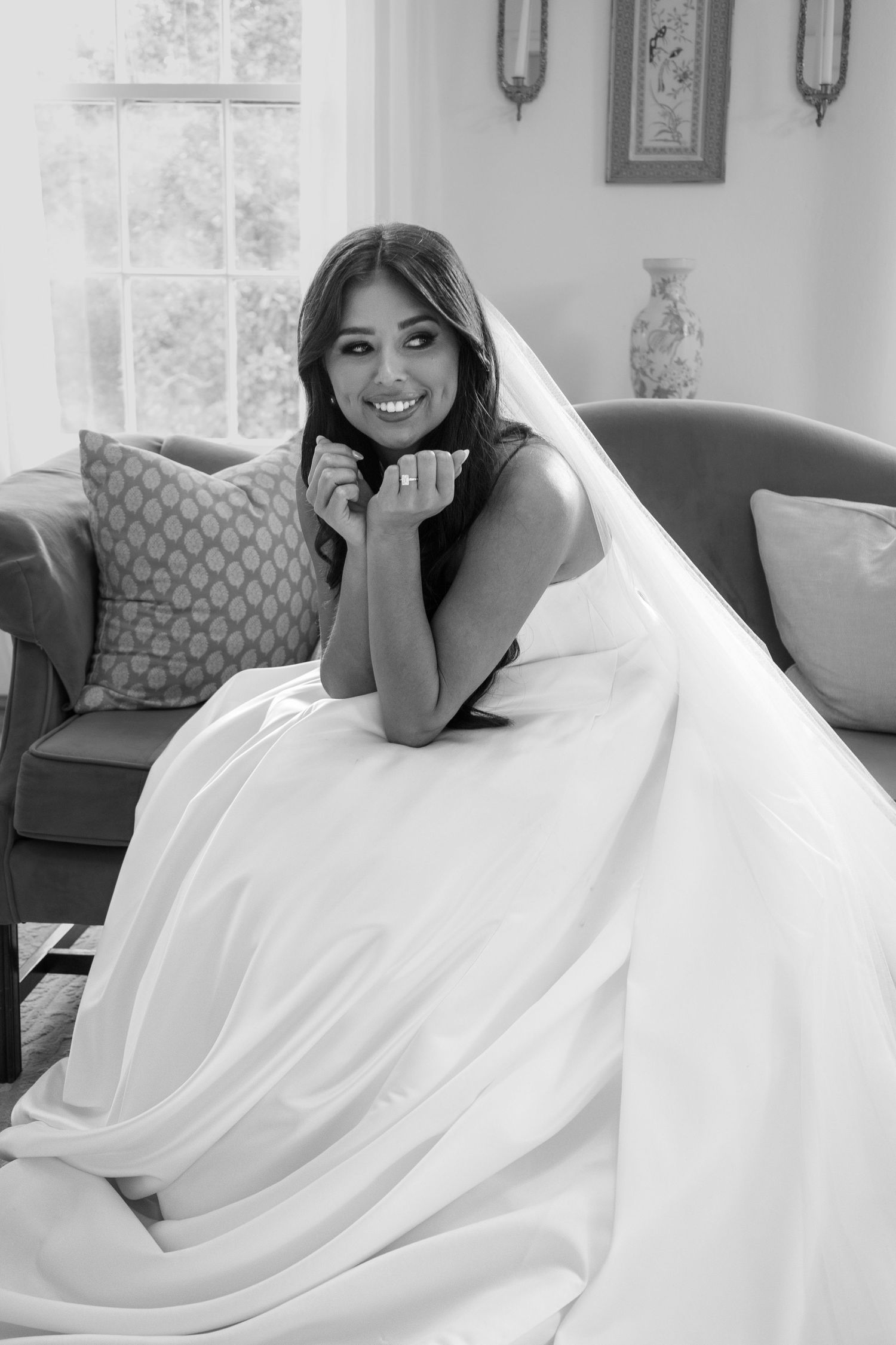 Black and white photo of a smiling bride in a white gown and veil, posing elegantly on a sofa near a window.