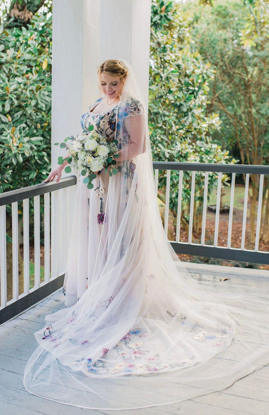 Bride in floral embroidered gown with sheer cape holds white bouquet on porch surrounded by lush greenery.