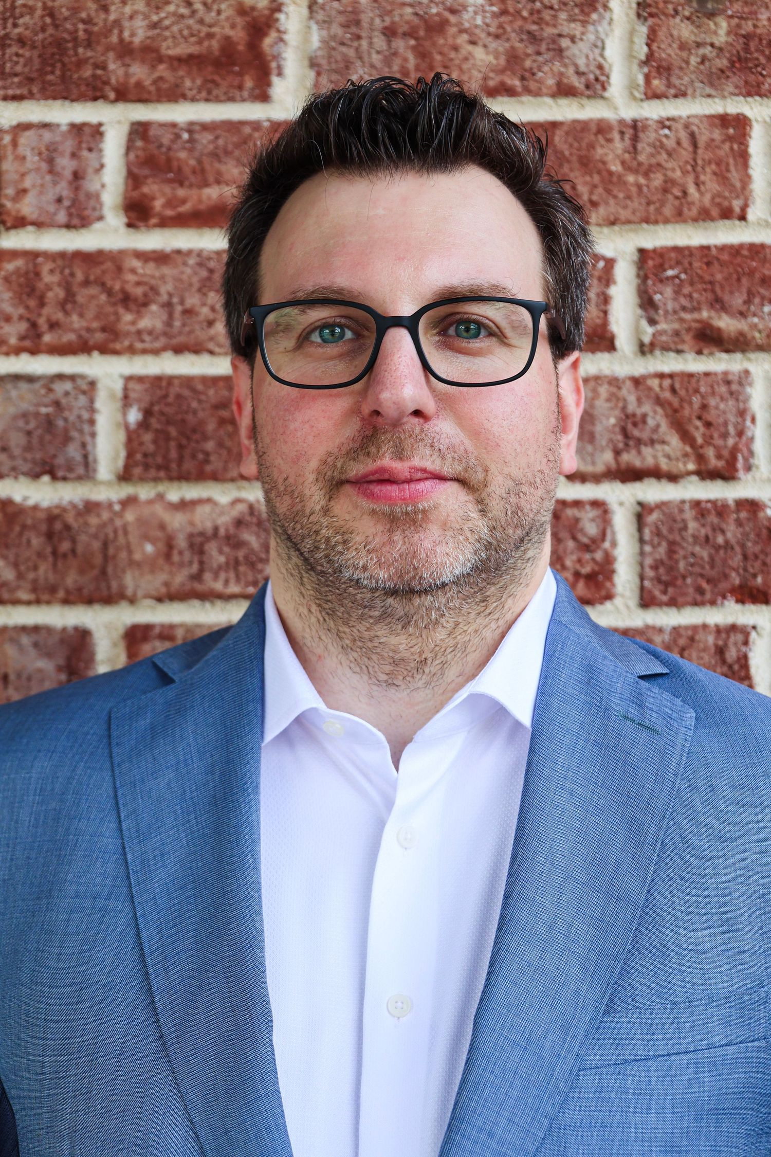 Professional man in blue blazer and glasses posing against a red brick wall for a business headshot.