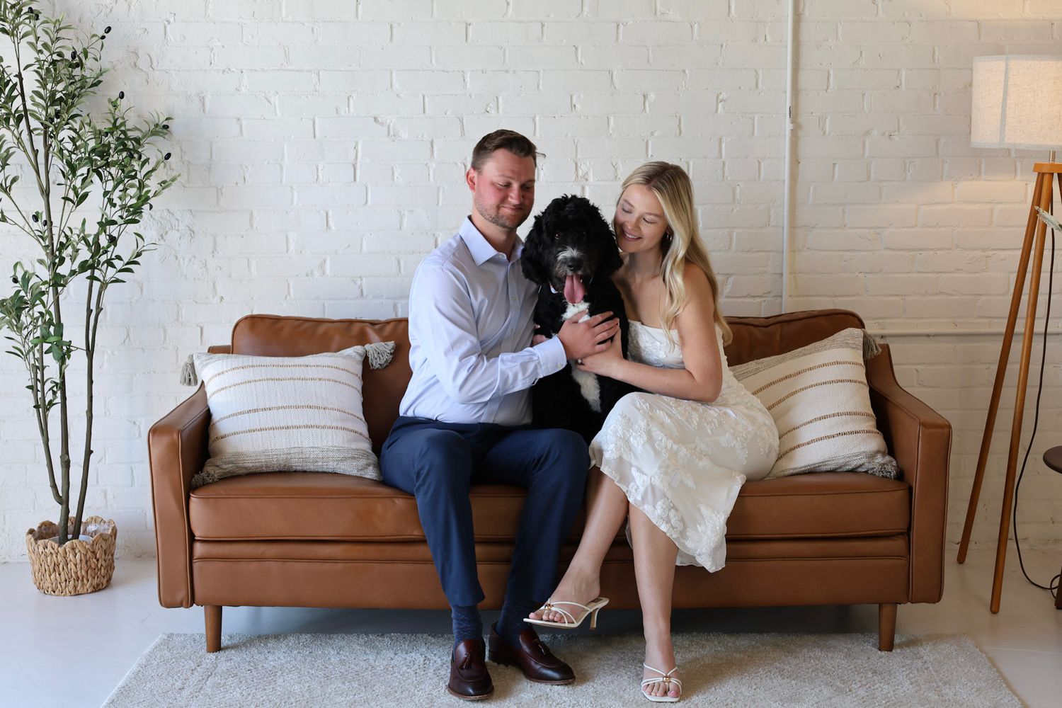 Couple sitting on brown leather sofa with black dog in bright white brick room with indoor plant and floor lamp.