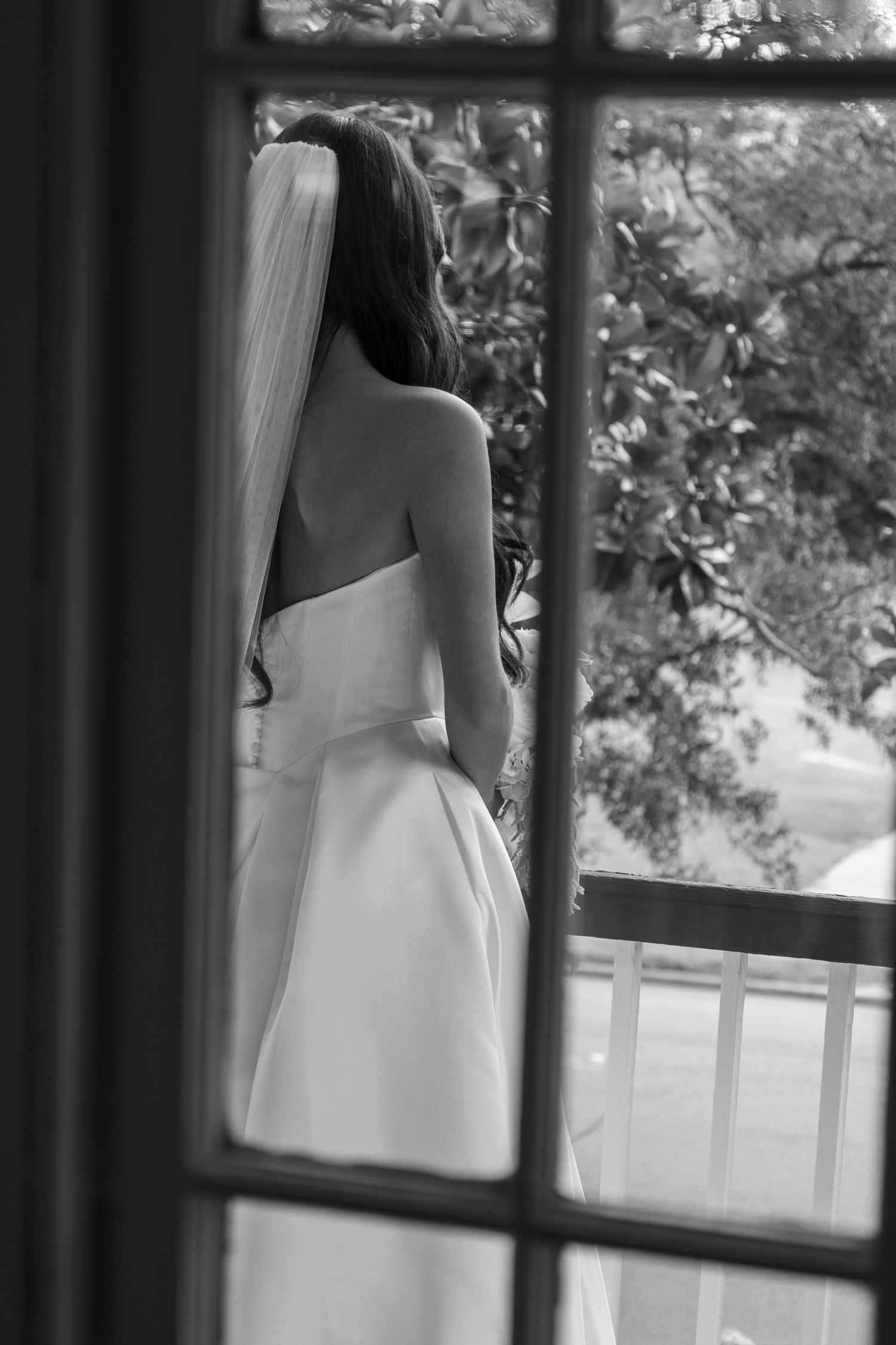 Black and white photo of a bride in a strapless gown and veil, viewed from behind through a window on a balcony.