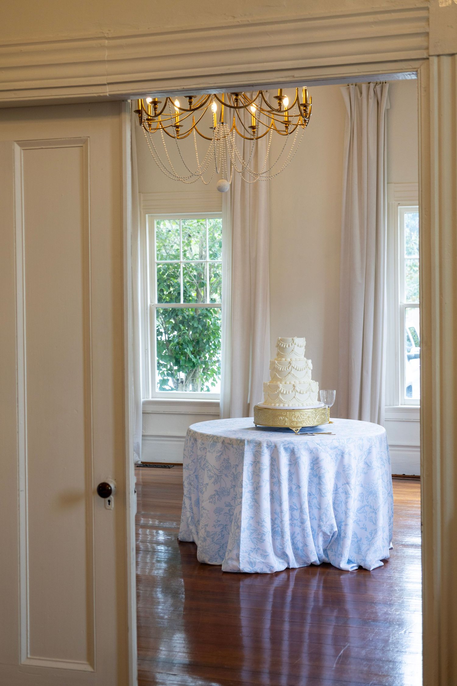Elegant white wedding cake on a draped table beneath a gold chandelier in a bright, classic reception room.