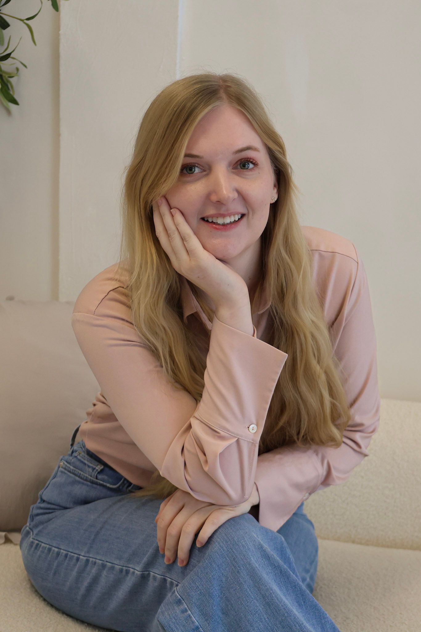Smiling blonde woman in pink shirt and jeans sitting on sofa with hand on cheek in bright room.