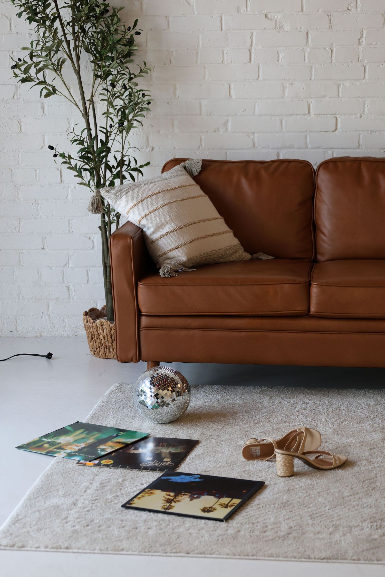 Brown leather sofa with striped pillow, disco ball, vinyl records, and sandals on a cream rug by a white brick wall.