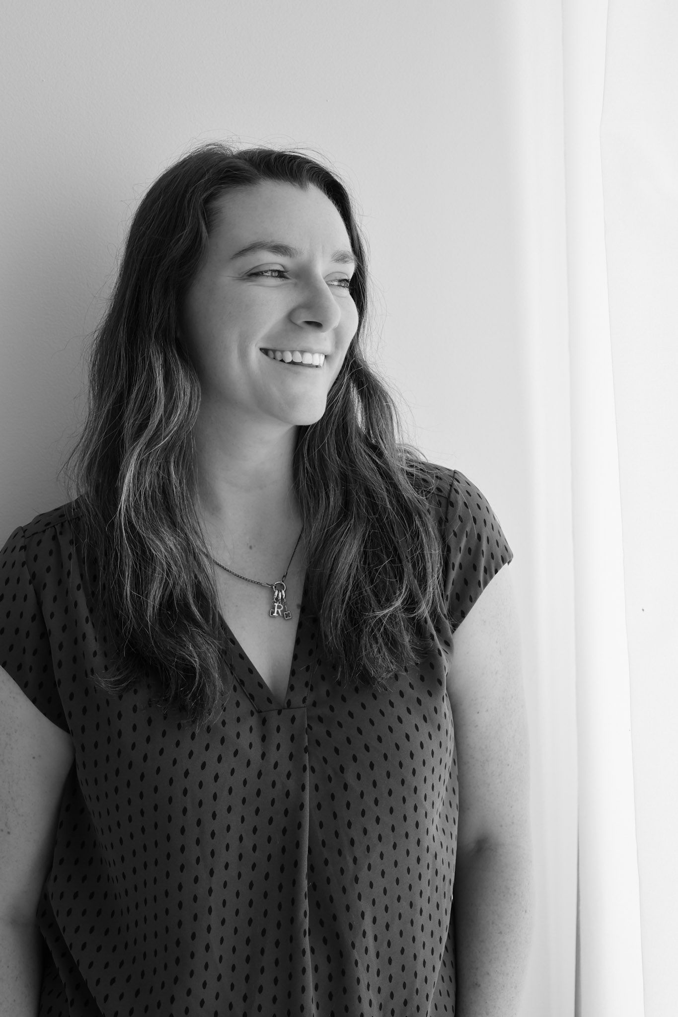 Black and white portrait of smiling brunette woman in polka dot blouse standing by window.