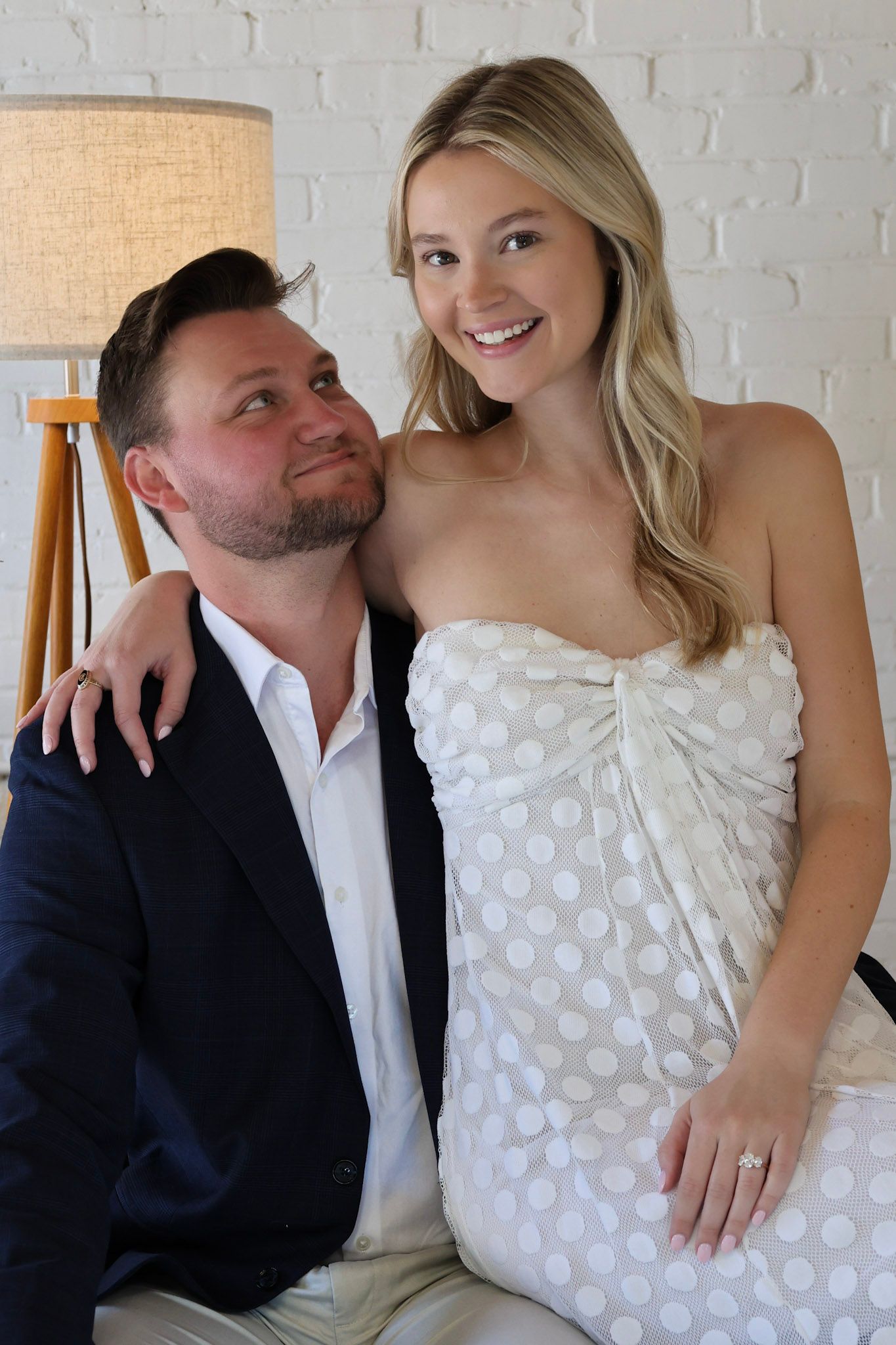 A smiling couple poses together, woman in white polka dot dress with engagement ring, man in navy suit jacket.