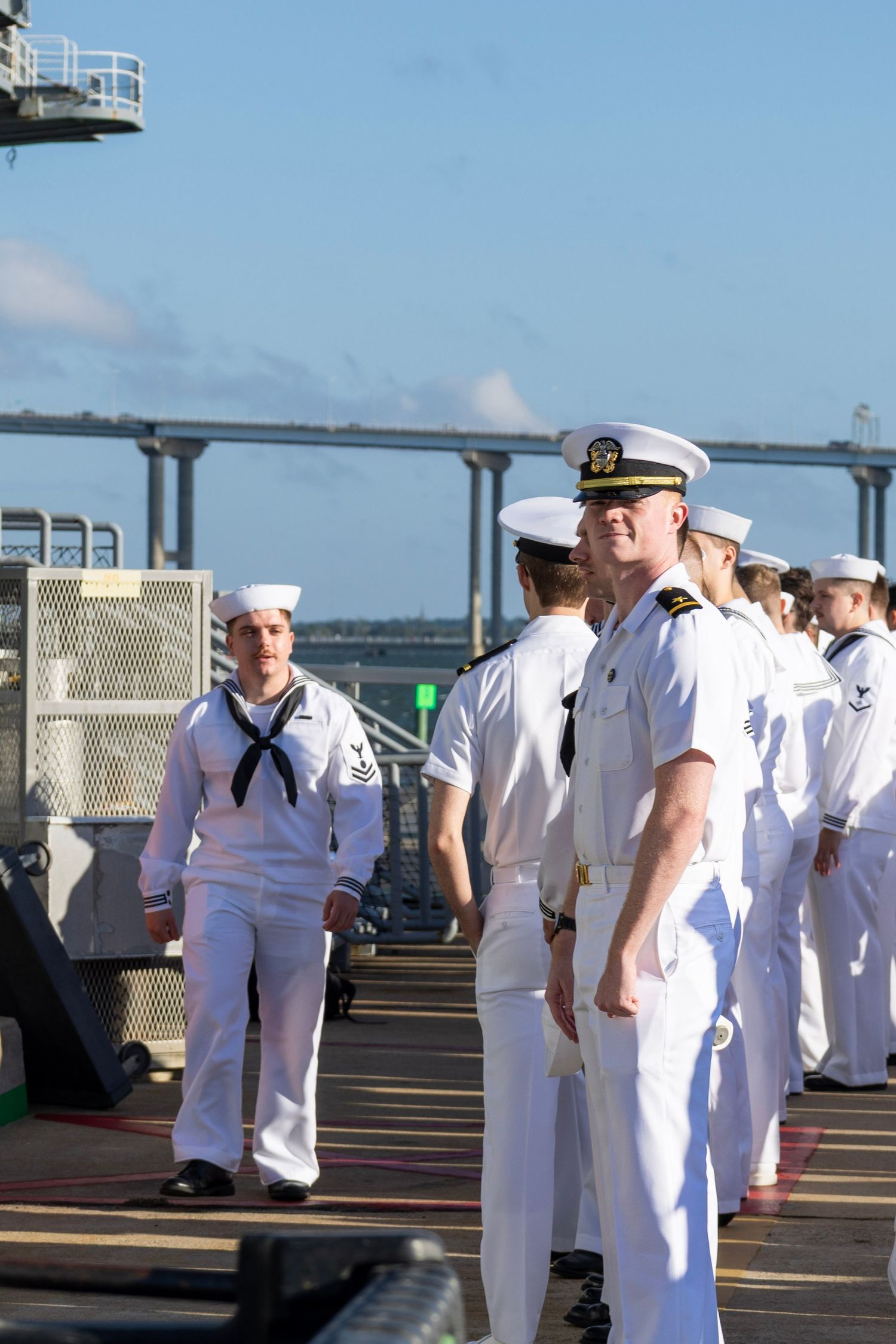 US Navy sailors in white dress uniforms stand at attention on a ship deck with a bridge visible in the background.