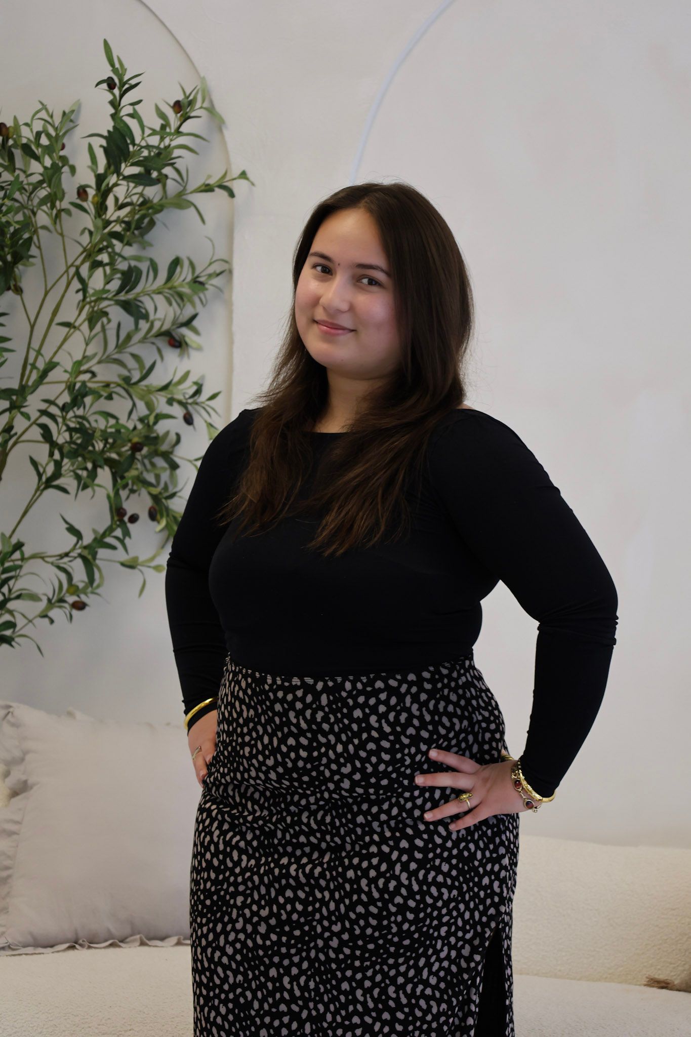 Young woman in black top and leopard print skirt posing indoors with green plant decor in background.