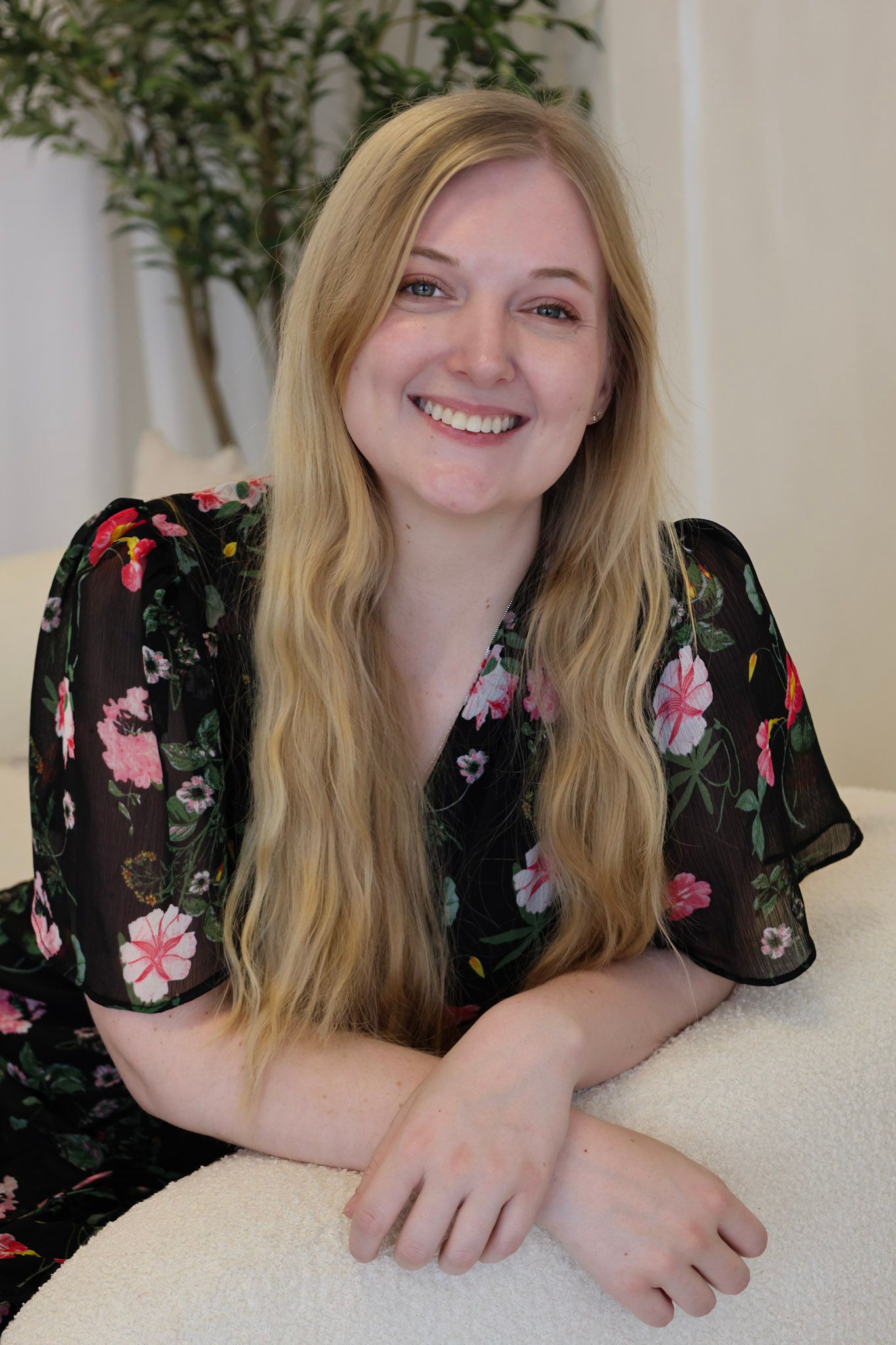 Smiling young woman with long blonde hair wearing a black floral dress, posing casually on a white couch indoors.