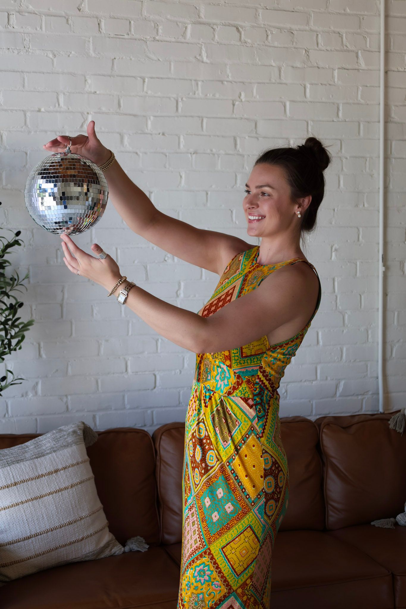 Woman in colorful patterned dress holding a disco ball in a modern living room with white brick wall.