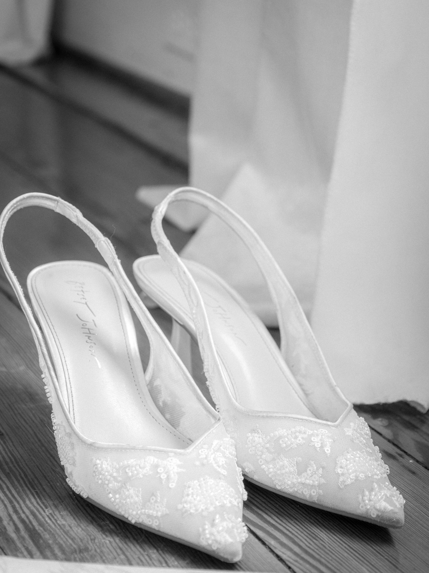 Elegant white lace slingback bridal heels with floral embroidery displayed on wooden floor in black and white photo.