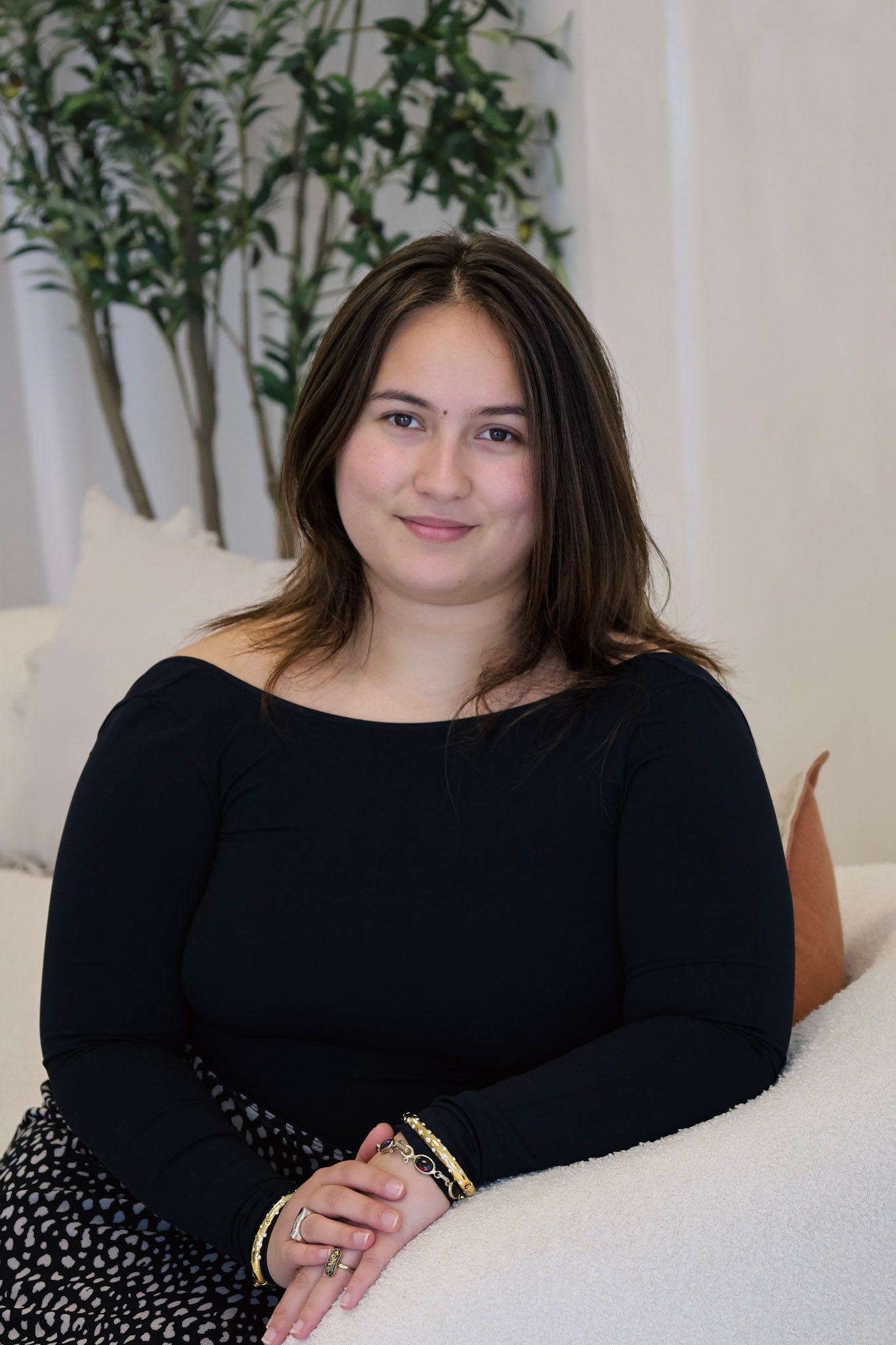 Young woman in black off-shoulder top sitting on white sofa, smiling warmly in bright modern interior.