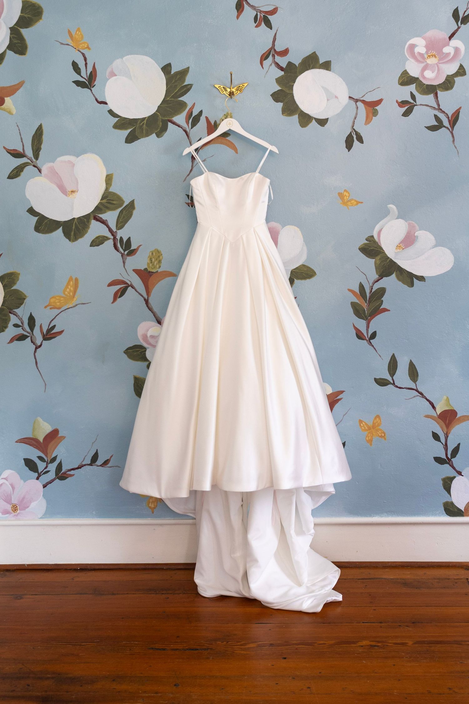 Elegant white satin wedding gown with sweetheart neckline hanging on a hanger against a blue floral wallpaper backdrop.
