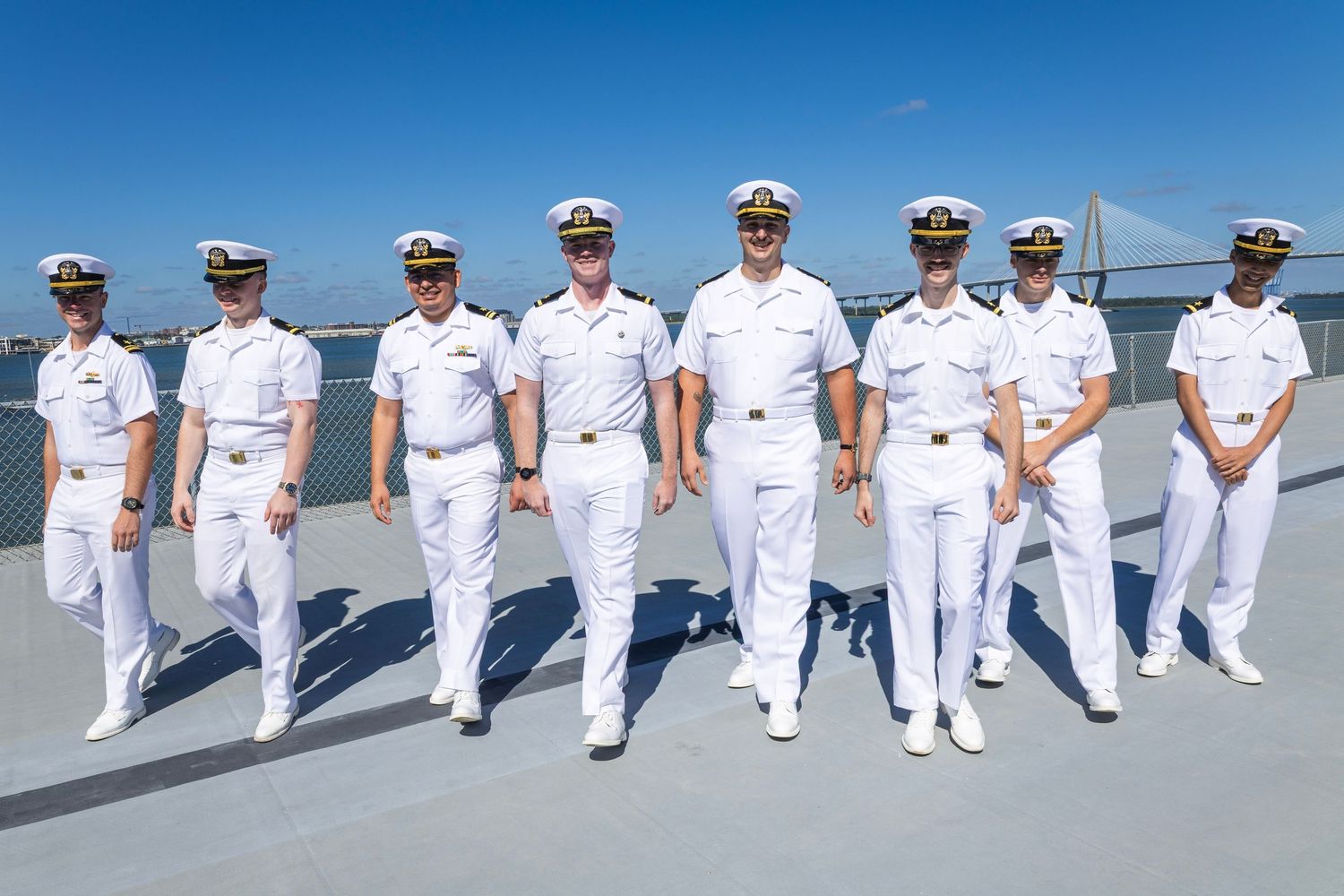 Eight Navy officers in white dress uniforms pose together on a ship deck with a suspension bridge visible in the background.