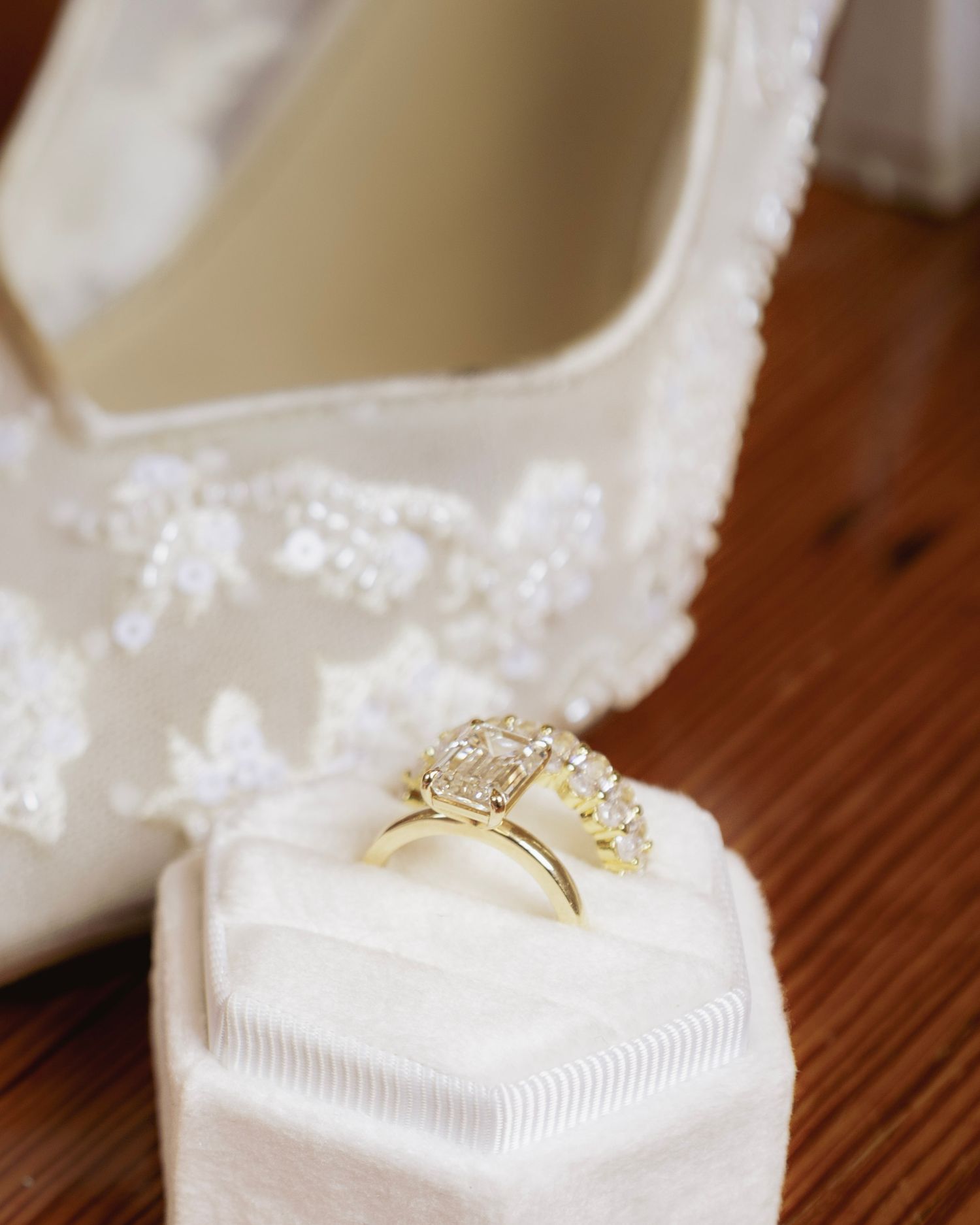 Gold diamond engagement ring and wedding band displayed on white velvet ring box beside lace bridal heels.