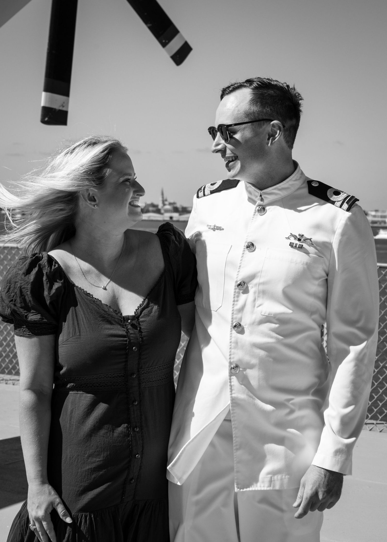 Black and white photo of a smiling couple, woman in dress and man in white naval uniform, standing outdoors on a sunny day.
