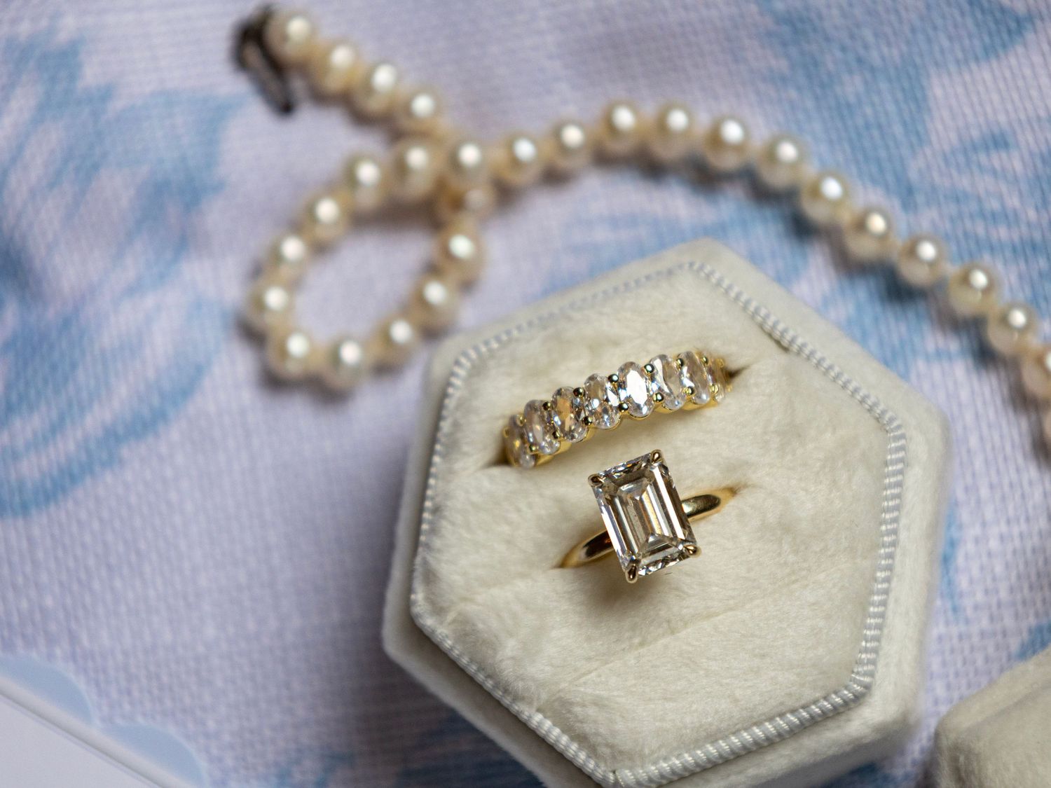 Gold diamond engagement ring with emerald cut stone in velvet box beside pearl necklace on blue floral fabric.