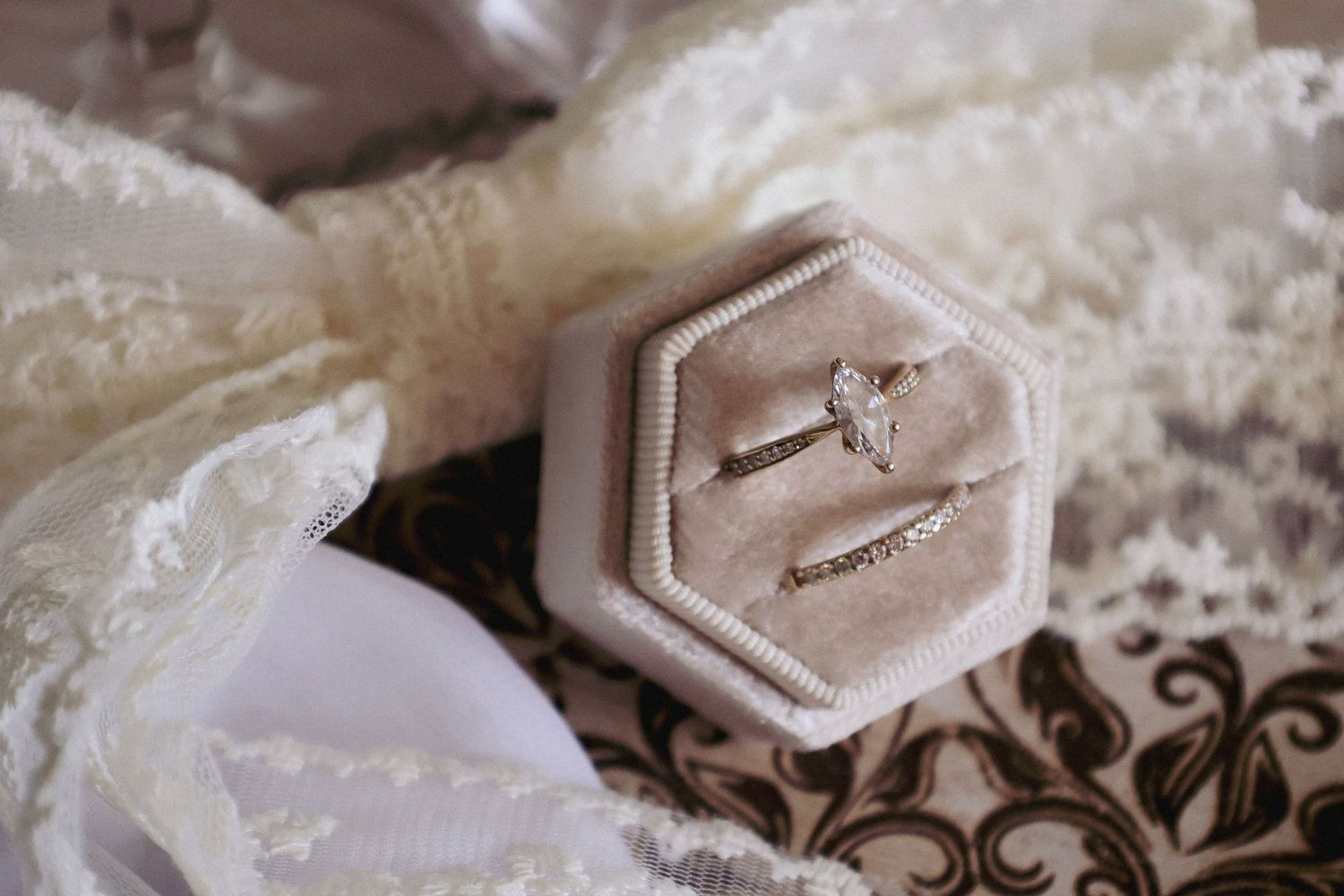 Diamond engagement ring and wedding band displayed in a hexagonal velvet ring box on lace fabric.
